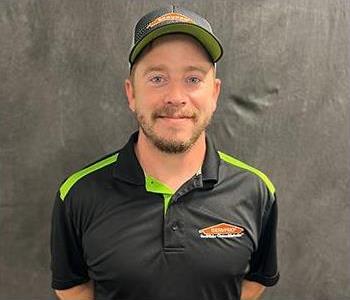 Male person grinning with short light brown beard with stunningly piercing blue eyes wearing a black shirt and ballcap.